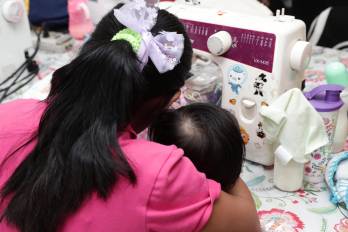 Una niña realiza un vestido mientras sostiene a su hijo en brazos.