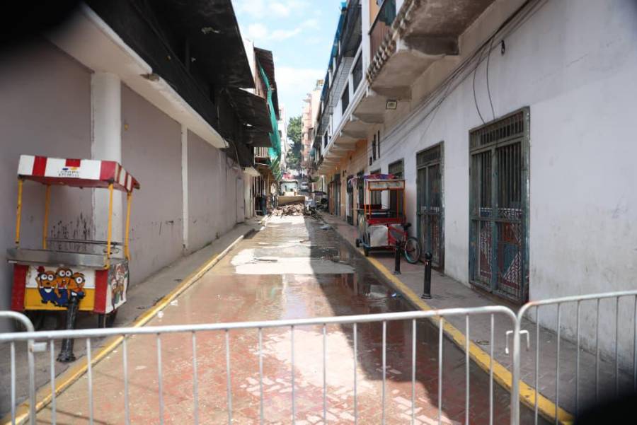 Vista longitudinal del callejón posterior al siniestro, bloqueado en primer plano por vallas metálicas de seguridad que impiden el acceso al público. El asfalto se encuentra notablemente húmedo debido a las labores de los bomberos y de limpieza. A los costados permanecen carritos de comida rápida inmóviles, mientras a lo lejos continúa el operativo de remoción